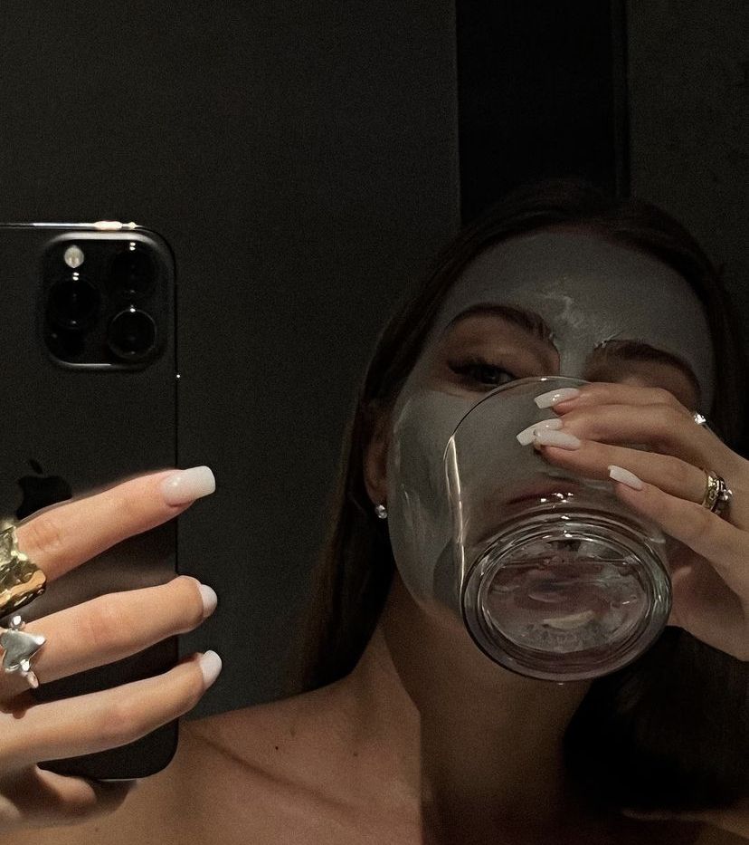 Person with mud mask holding a phone and a glass with a dark background